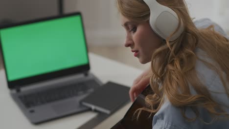 country female musician is playing guitar at home, sitting in front of open laptop with green screen, streaming online in video chat