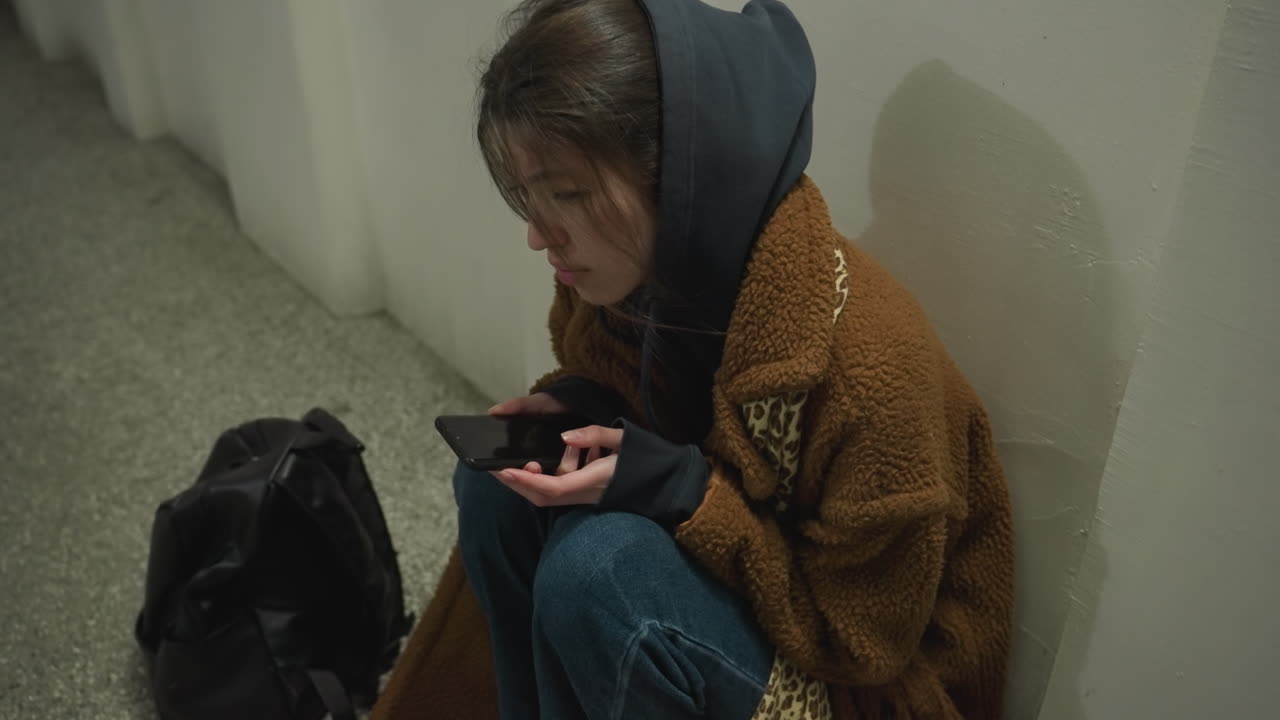 una chica con un abrigo marrón se sienta inclinada en un túnel subterráneo, sosteniendo su teléfono con una expresión de angustia. parece profundamente preocupada y perdida en sus pensamientos