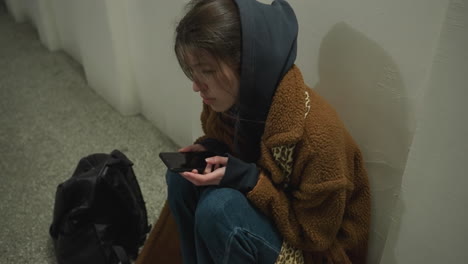 a girl in a brown coat sits bent over in an underpass tunnel, holding her phone with a distressed expression. she appears deeply troubled and lost in thought