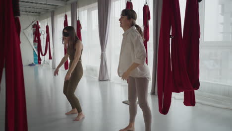 wellness advocate and trainee shake their bodies in rhythmic relief practice with blindfolds on hands loose under red silk hammocks in studio for tension release and mind body connection