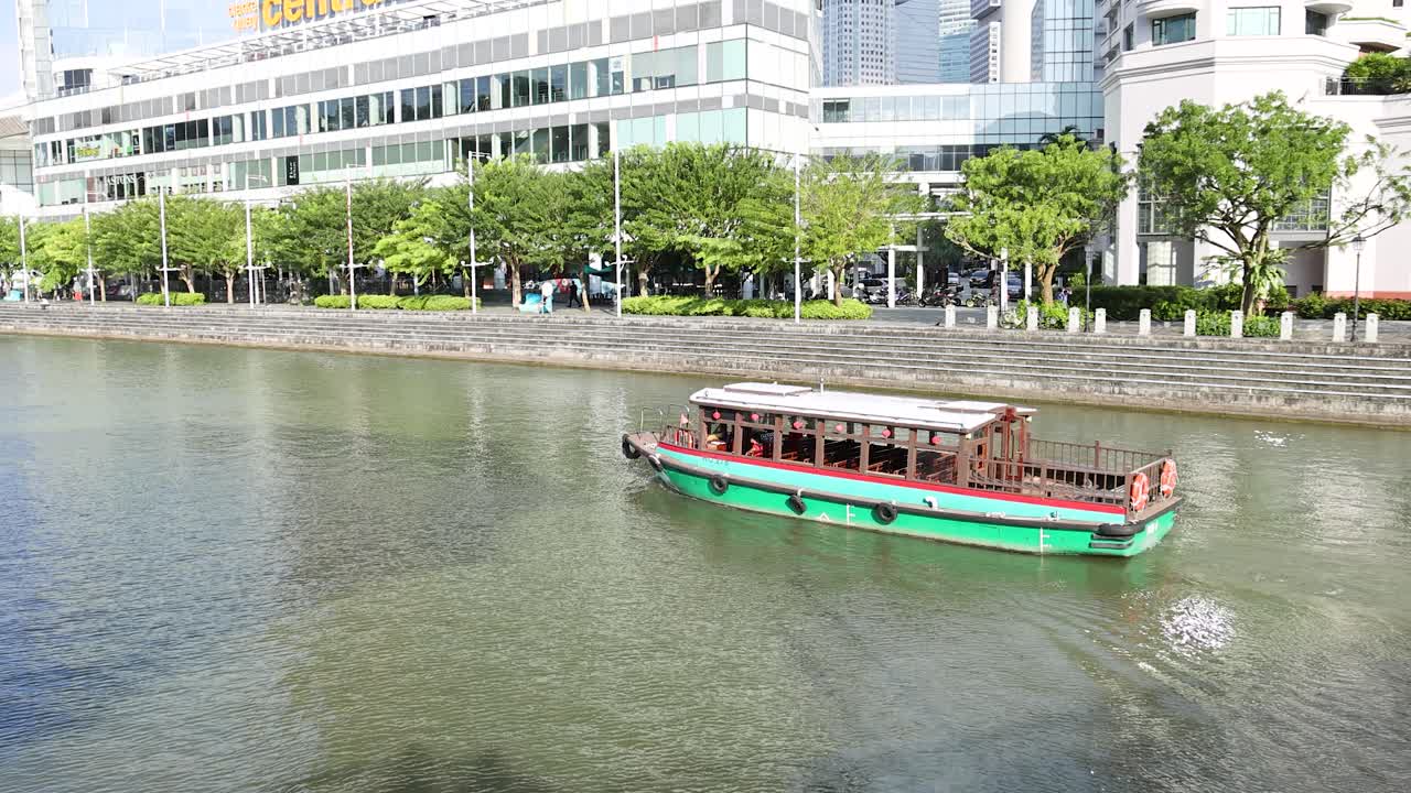 A traditional river cruise boat travels across a sunlit urban river, passing modern buildings and greenery in downtown Singapore. Wide, steady camera angle