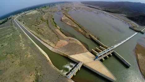 Top-Aerial-view-of-spillway-of-a-dam,-Beautiful-majestic-mountains-in-the-back-of-the-dam,-Sun-shines-in-the-green-water-of-the-dam