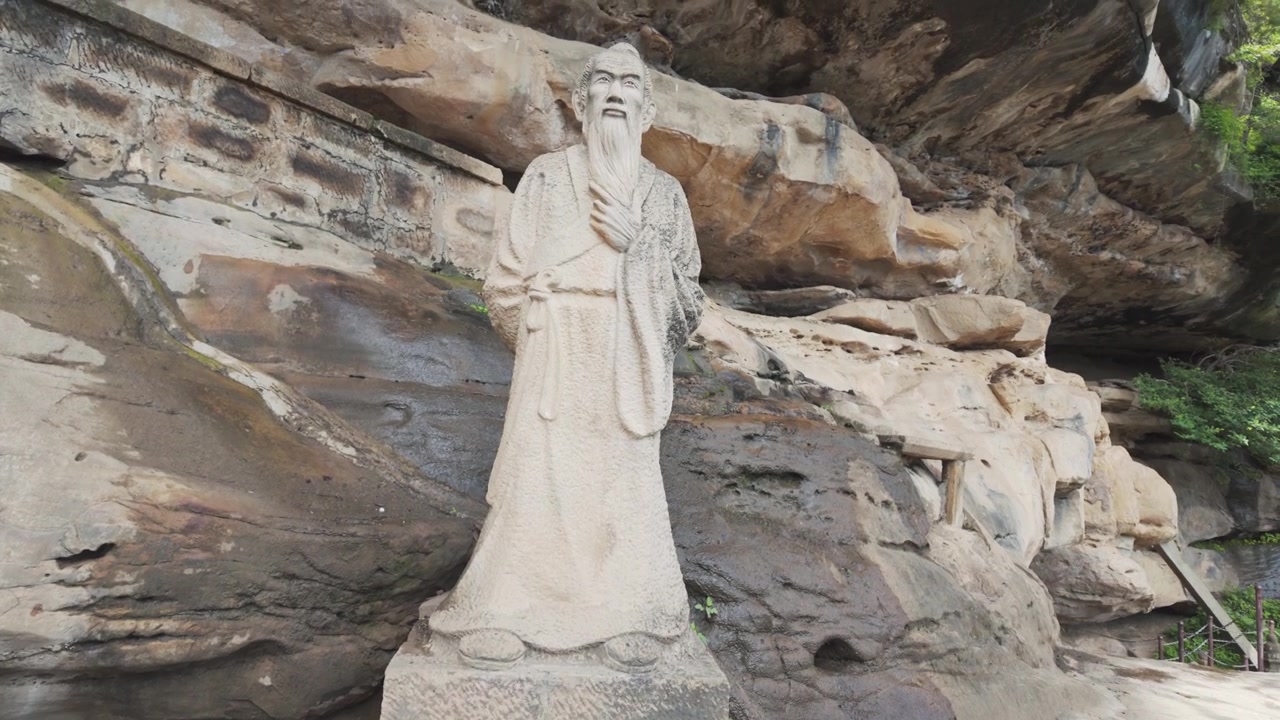 Una scultura in pietra dettagliata di un saggio o filosofo tradizionale cinese in piedi contro una scogliera scoscesa. Autentico patrimonio culturale e punto di riferimento storico.