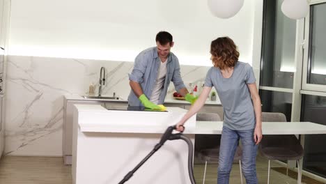 cleaning day. young couple cleaning their apartment - man in gloves wiping up kitchen stove, girl cleaning floor using vacuum cleaner. clean studio kitchen together in the evening. smiling to the camera