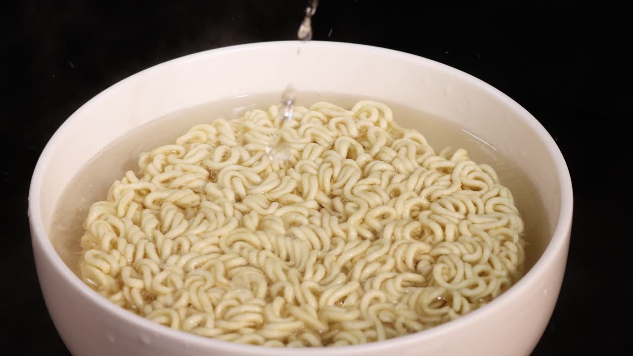 Hot water is steadily poured into a white bowl containing dry instant noodles, shot from above with even lighting and a static camera on a dark background