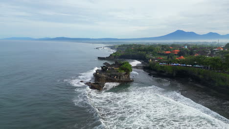 drone pushes up past tanah lot temple on a misty morning as waves roll in