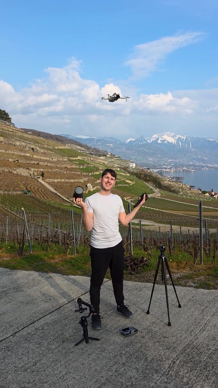Scatto verticale di un fotografo che pilota un drone nei vigneti svizzeri, con attrezzatura fotografica, treppiede, vista lago, colline terrazzate e montagne innevate sullo sfondo