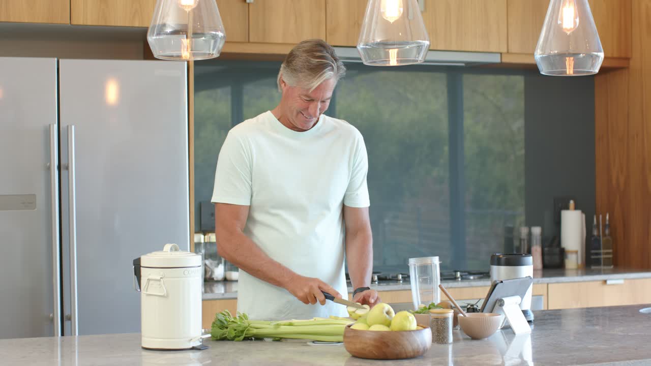 Älterer Mann folgt Tablet-Rezept, schneidet Apfel mit Messer an Kücheninsel und bereitet einen Snack zu.