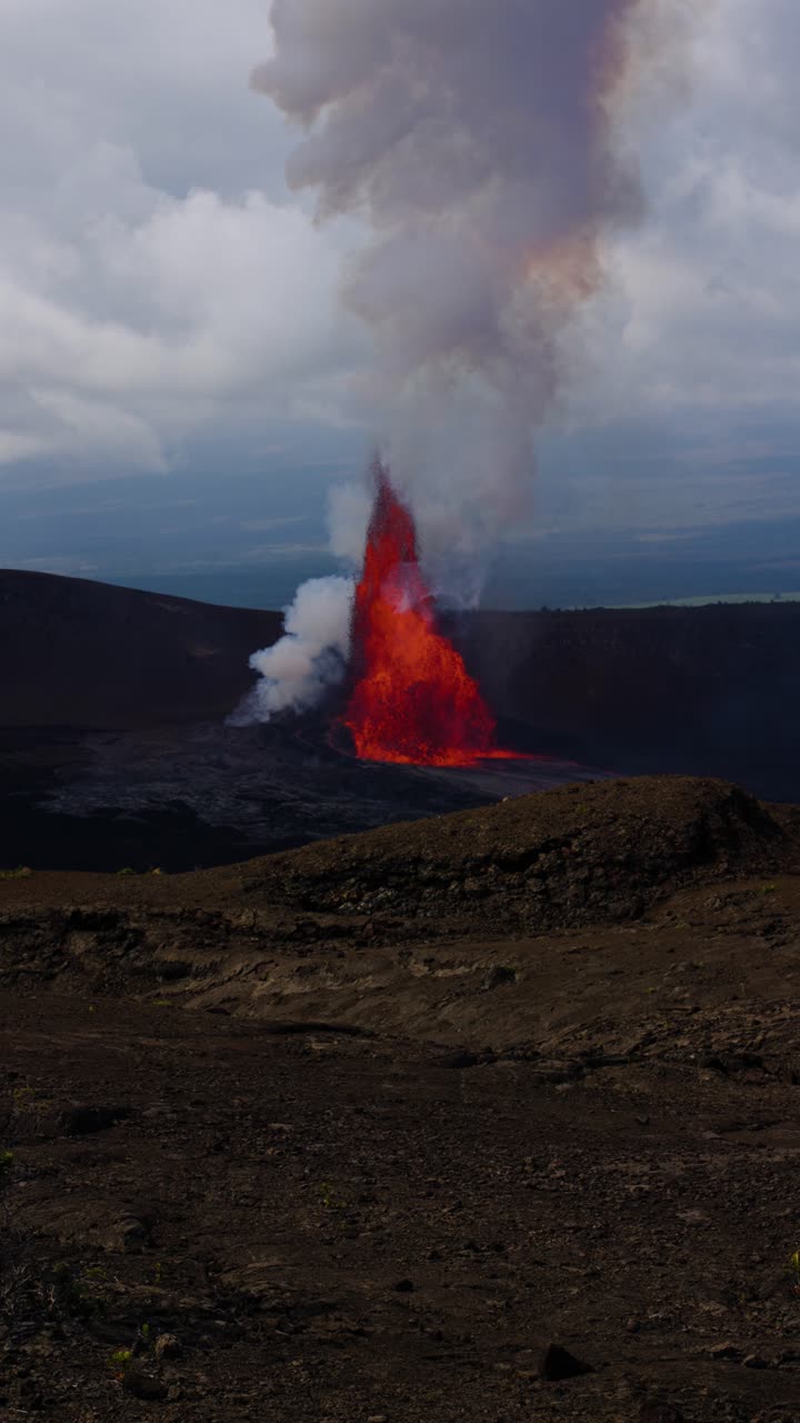 Inquadratura verticale statica dell'eruzione del Kīlauea sull'Isola Grande delle Hawaii, che mostra un'intensa fontana di lava e un denso pennacchio di fumo che si innalza sopra il cratere vulcanico.
