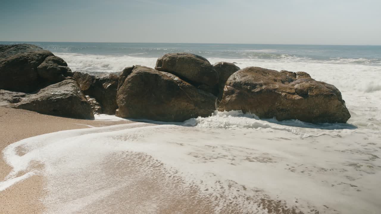 Sunlit ocean waves wash around large coastal rocks on a sandy beach, creating white sea foam in a serene summer seascape with natural textures and copy space