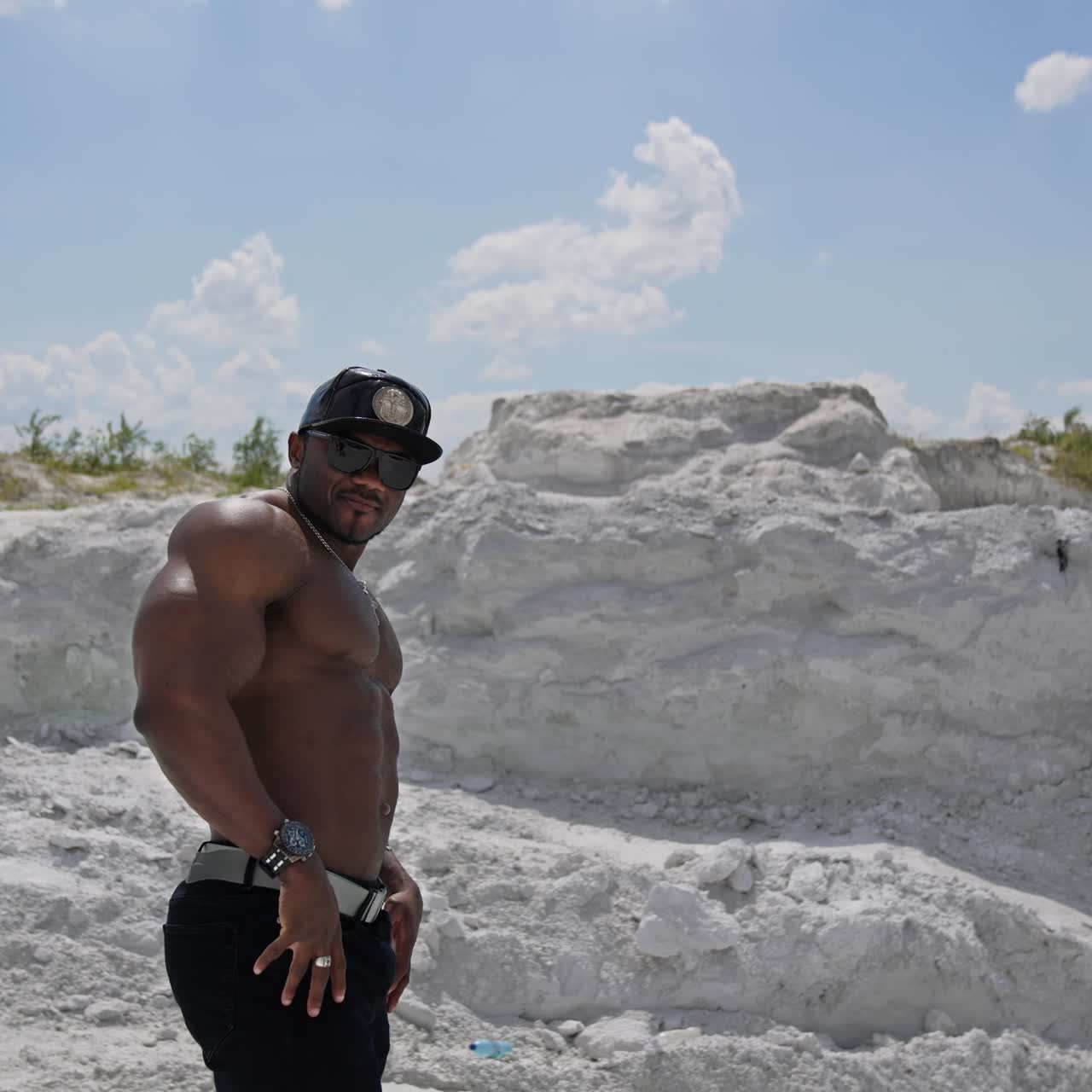 Muscular athlete posing on camera. Dark skinned sportsman in black cap and sunglasses showing his trained body on white rocky background. Circling view