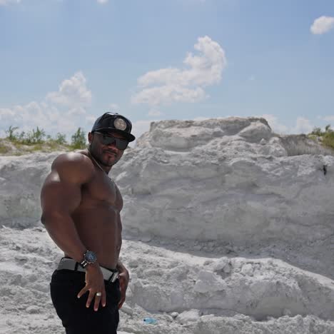 muscular athlete posing on camera. dark skinned sportsman in black cap and sunglasses showing his trained body on white rocky background. circling view