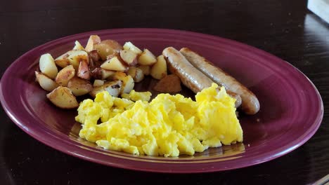 Maroon-plate-with-cooked-breakfast-sausage,-potatoes-and-scrambled-eggs-steaming