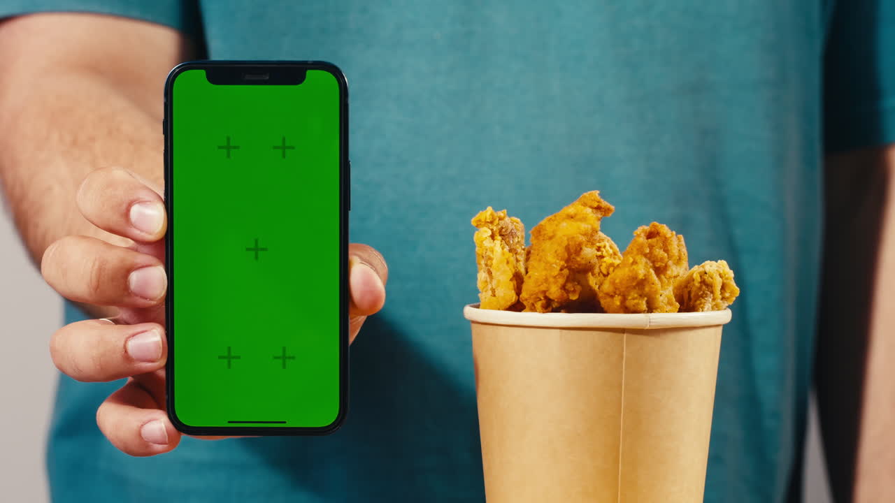 Person holding a green screen smartphone next to a container of fried chicken