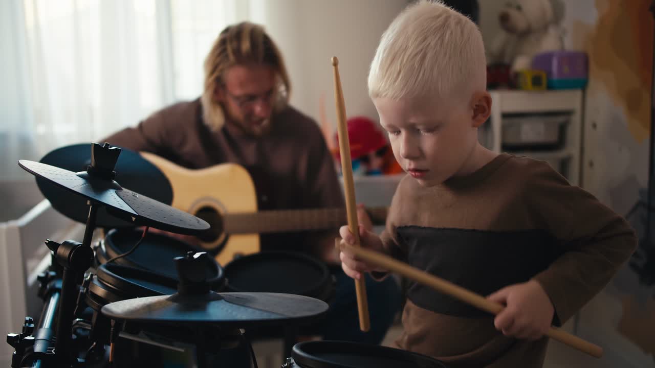 Happy albino boy with white hair energetically plays the drum set with the help of special sticks and his dad, a blond man with a beard and glasses, plays an acoustic guitar with his son in a room for developing talents for playing musical instruments at home
