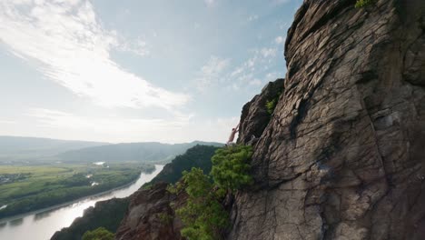 aerial-of-mountain-climber,-man-bouldering,-rocky-mountaintop,-guy-free-climbs-wall,-rope-climbing-in-Austria,-Durnstein,-sunny-drone-shot,-europe-outdoor-sports-travel,-flying-FPV,-river-Danube