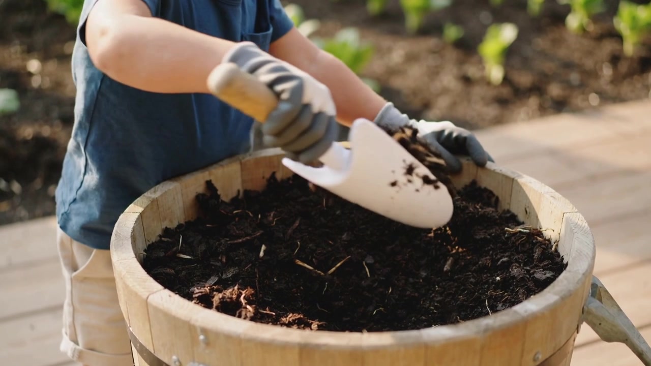 Un bambino si sporge su un barile in un patio pavimentato, raccogliendo terra con una paletta e guanti, preparando il terreno.
