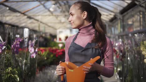 joven jardinera en uniforme caminando en un invernadero entre estantes con diferentes plantas y sosteniendo un riego de naranja en sus manos