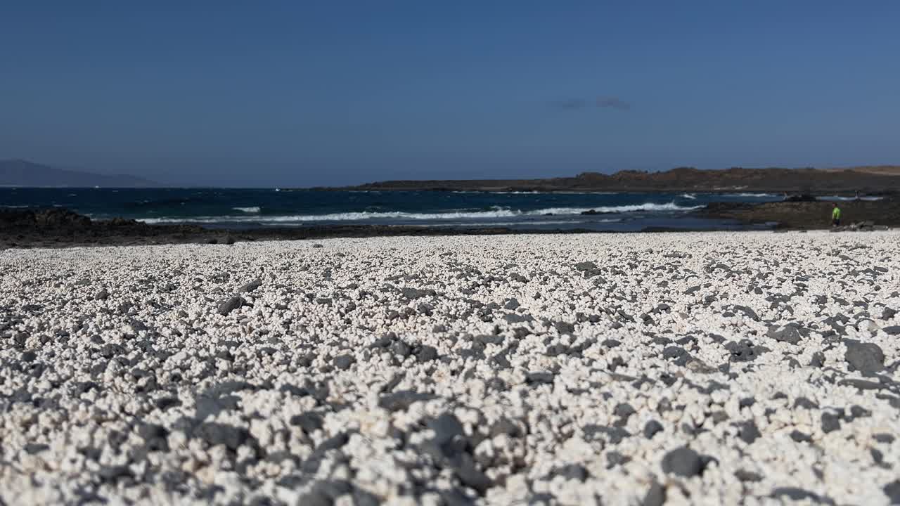 Low-angle static shot focusing on unique white rhodoliths of Popcorn Beach, with the Atlantic Ocean and a distant person in the background. Fuerteventura, Spain