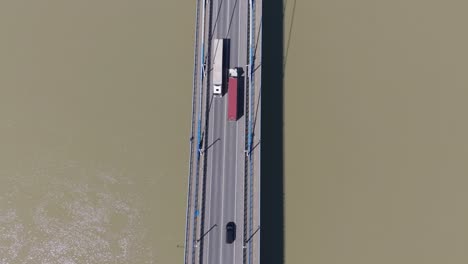 an aerial view of a cable-stayed bridge shows cars and trucks traveling over a wide river. the straight road and vehicle shadows emphasize the structure’s geometry and the movement across the water