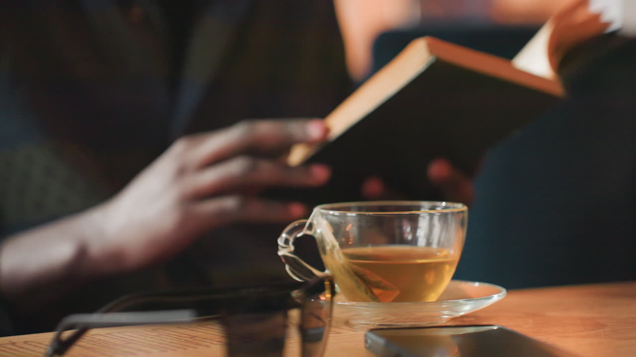 찻잔 옆에서 책 페이지를 넘기는 손가락의 클로즈업, 찻잔이 놓인 근처에서 손으로 페이지를 넘기는 디테일 샷, 나무 표면의 따뜻한 차 옆에서 손가락으로 페이지를 넘기는 친밀한 장면