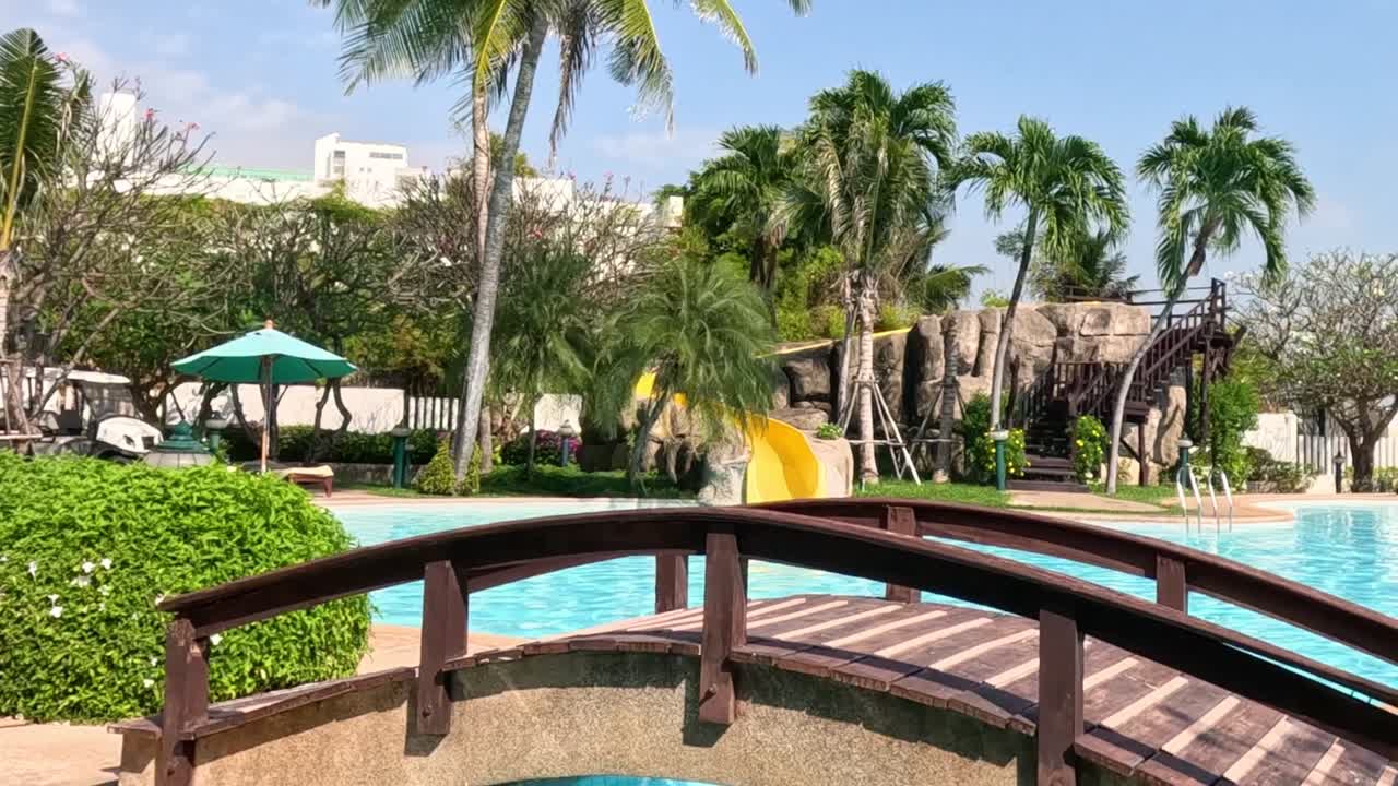 A scenic view of a wooden bridge with lush palm trees and a bright poolside setting.