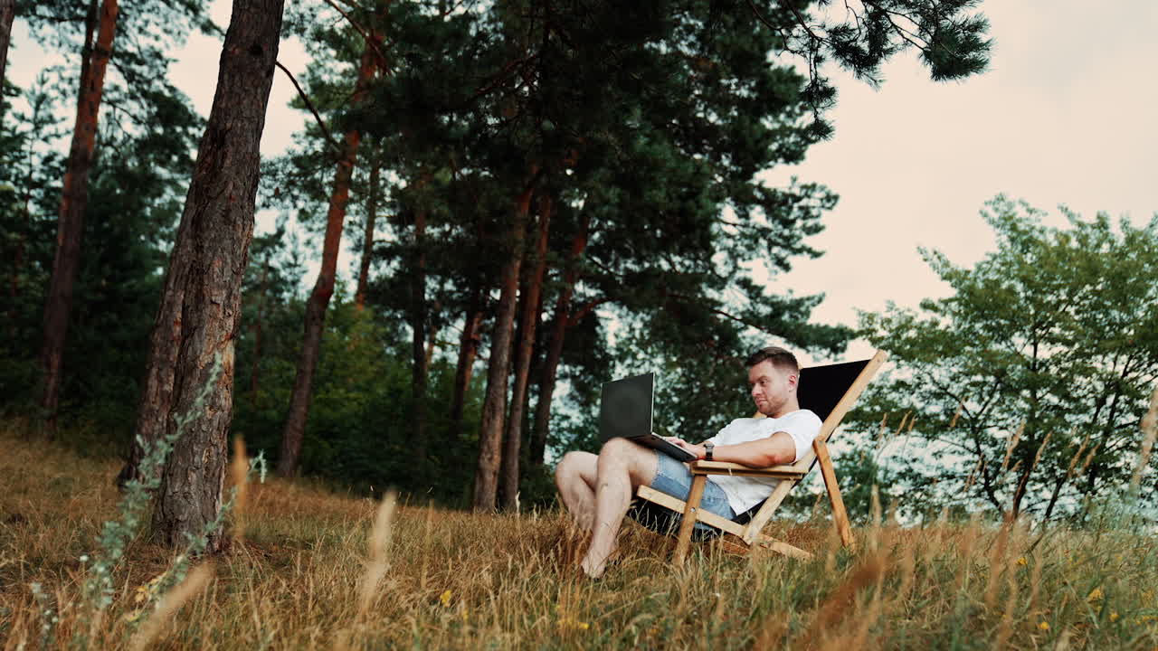 Approaching a man sitting in the folding chair. Freelancer works on laptop remote. Low angle view. Nature at backdrop.