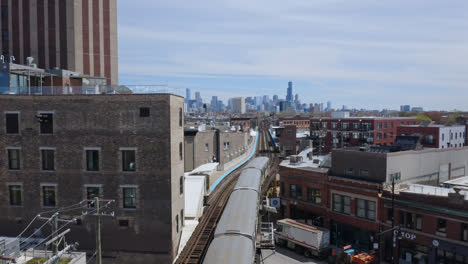 long gray subway slows down on the elevated track between modern and traditional buildings to enter damen station with the chicago skyline in the background