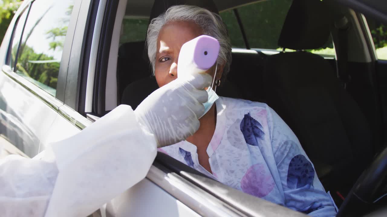 trabajador de la salud con ropa de protección midiendo la temperatura de una mujer mayor afroamericana en un coche