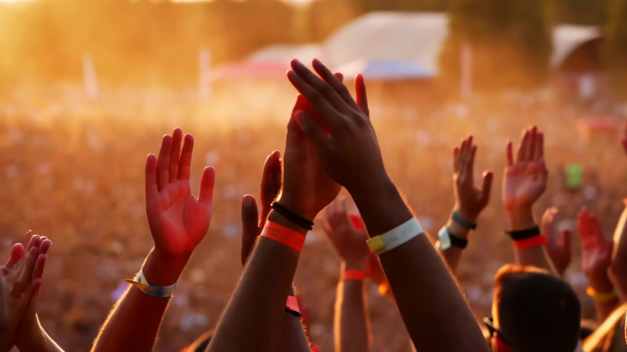 Close foreground view shows hands repeatedly coming together and separating above packed gathering, while orange backlight and soft haze silhouette arms against blurred crowd.