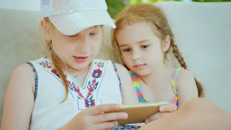 Two-Little-Girls-Are-Playing-On-The-Smartphone-They-Sit-Next-To-Each-Other-On-The-Beach-Chaise-Longu