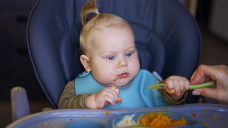 little infant baby sitting in high chair pulls the spoon from his mom. cute kid holds the spoon and doesn't let go.