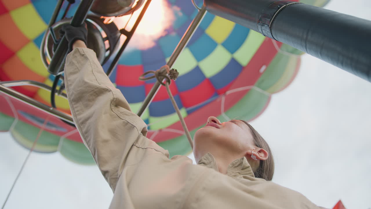 Gloved woman reaches up operating hot air balloon burner lever while vibrant patchwork envelope fills sky overhead, teammates gaze upward listening to roar of flame during serene sunrise ascent