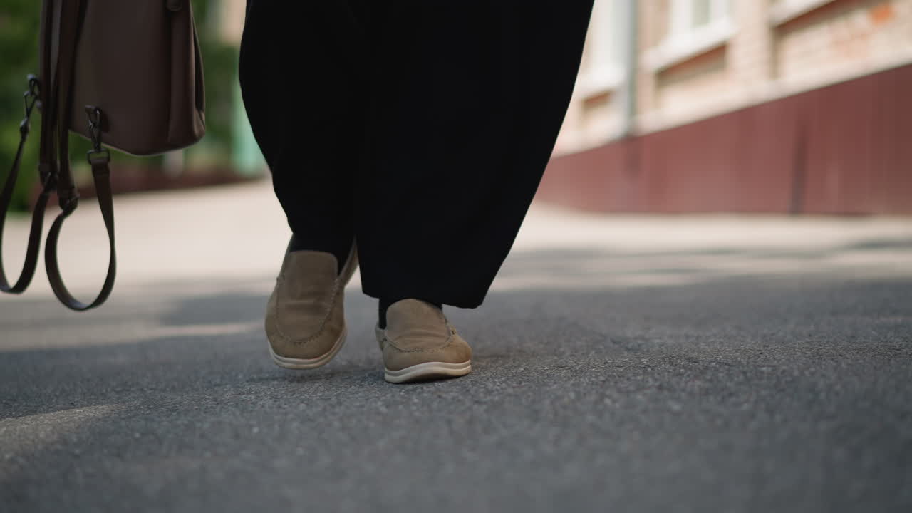 Leg view of girl walking outdoors on sunny day carrying brown handbag with strap gently touching pavement, sunlight casting soft shadows