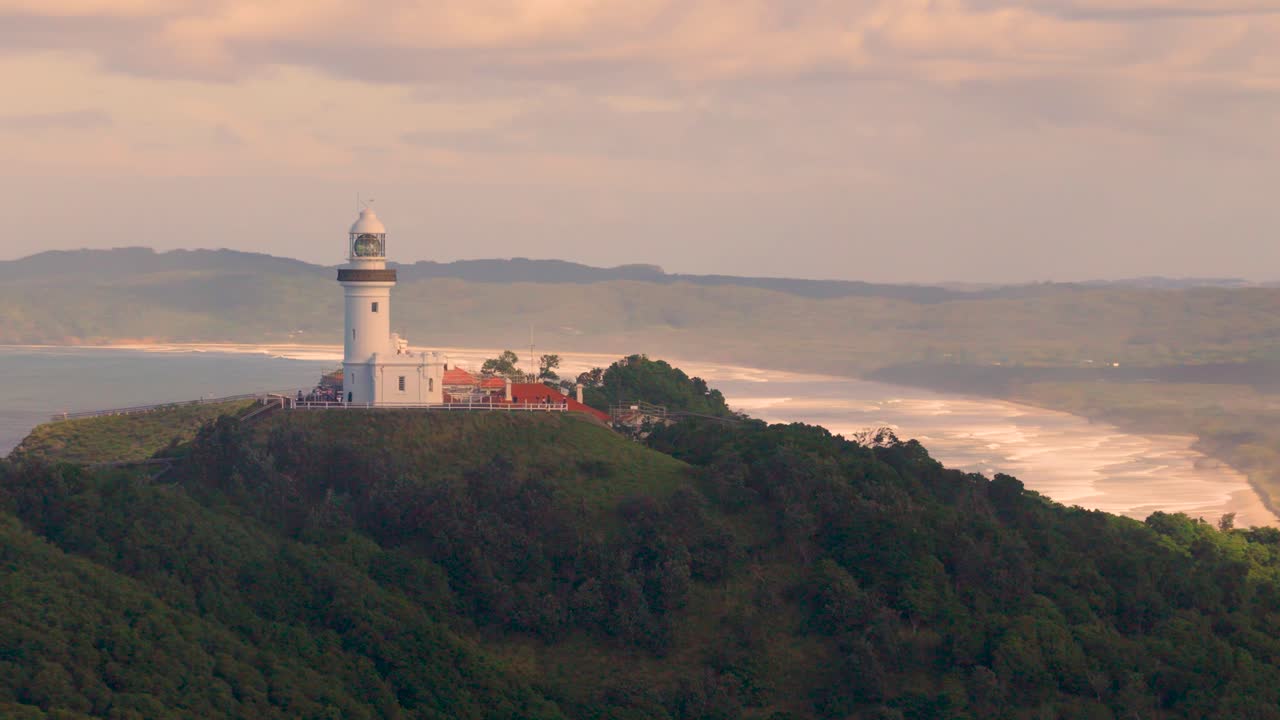 Drone footage captures the serene Byron Bay lighthouse atop lush hills with ocean views under soft evening light