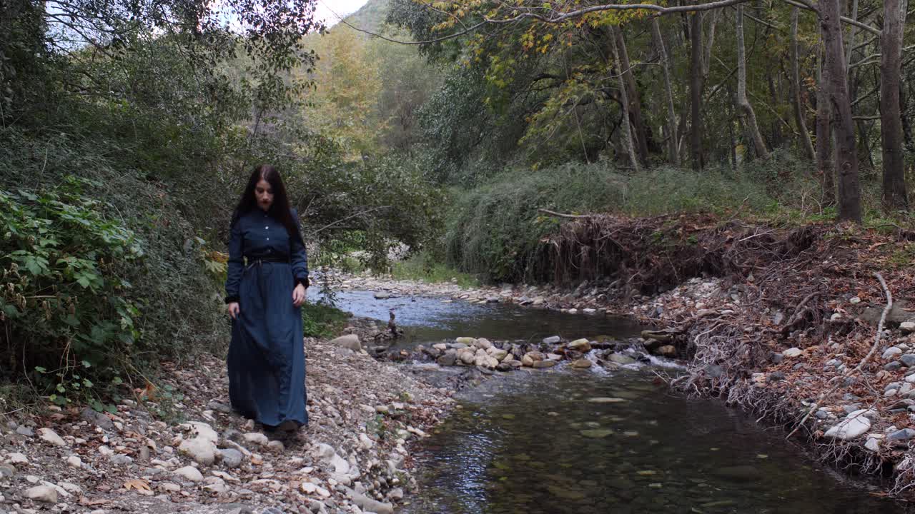 una mujer hermosa con un vestido gótico vintage camina sola junto a un río en el bosque