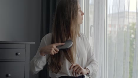 Dreaming-woman-brushing-brown-hair,-gazing-wistfully-out-the-window