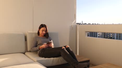 Young-woman-casually-lying-on-a-lounger-on-the-sunny-balcony-swiping-through-her-smartphone---Full-shot