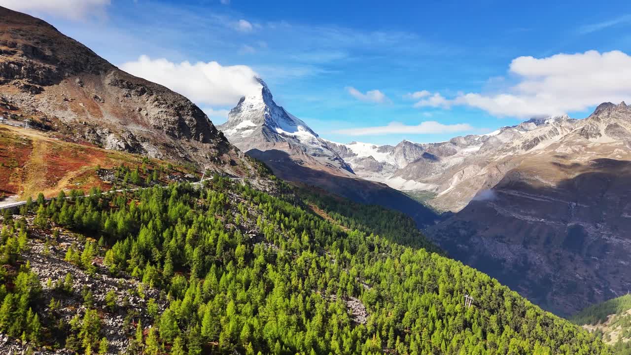 Aerial view of the iconic Matterhorn above Zermatt, with the Five Lakes Trail winding through autumn-colored larch forests and rocky alpine slopes in the foreground, under a vivid blue sky