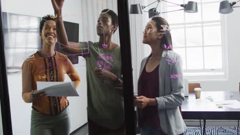 Diverse-businesswomen-standing-at-glassboard,-brainstorming-in-office