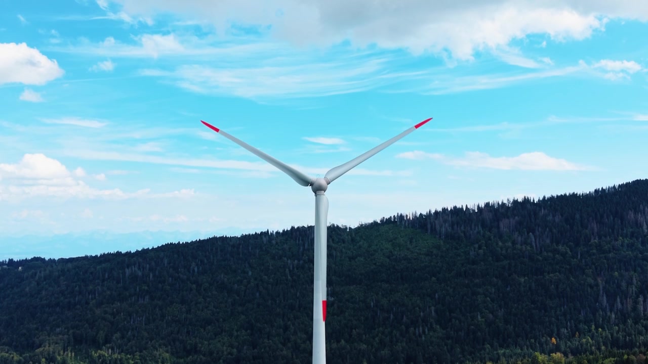 Wind turbine rotor and blades spin against wooded hills and wispy clouds in open blue sky, aerial descend tilt upward