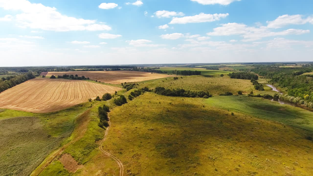 Green meadows, river, forests and wheat farmlands against blue sky. Shadows from clouds cover the ground.