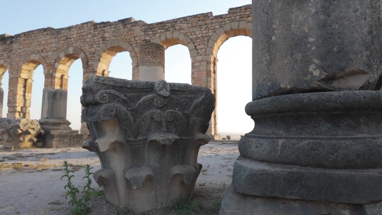 Cinematografisch beeld op grondniveau van een gebeeldhouwd Korinthisch kapiteel bij de Basiliek van Volubilis, dat Romeins architectonisch vakmanschap benadrukt op de archeologische UNESCO Werelderfgoedsite van Marokko.