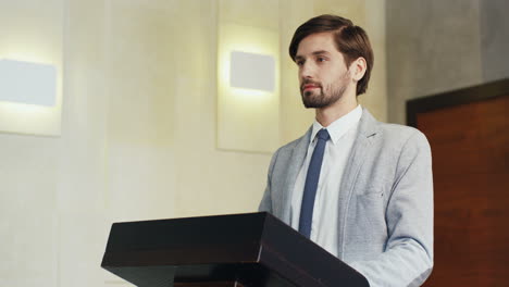 vista de primer plano joven hombre de negocios caucásico con ropa formal en un podio mirando a la cámara y sonriendo en una sala de conferencias