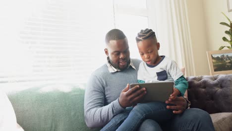 Happy-african-american-father-sitting-on-sofa-with-son-on-lap-using-tablet,-in-slow-motion