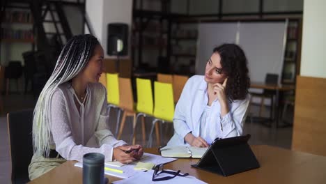 Dos-Mujeres-Atractivas-Sonriendo,-Conversando-En-Una-Biblioteca-Con-Estantes-De-Libros-En-Segundo-Plano-1