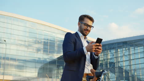 Attractive-man-wearing-glasses,-laughinging-while-texting-a-message-on-the-smartphone,-standing-with-a-bicycle-near-a-huge-glass-building