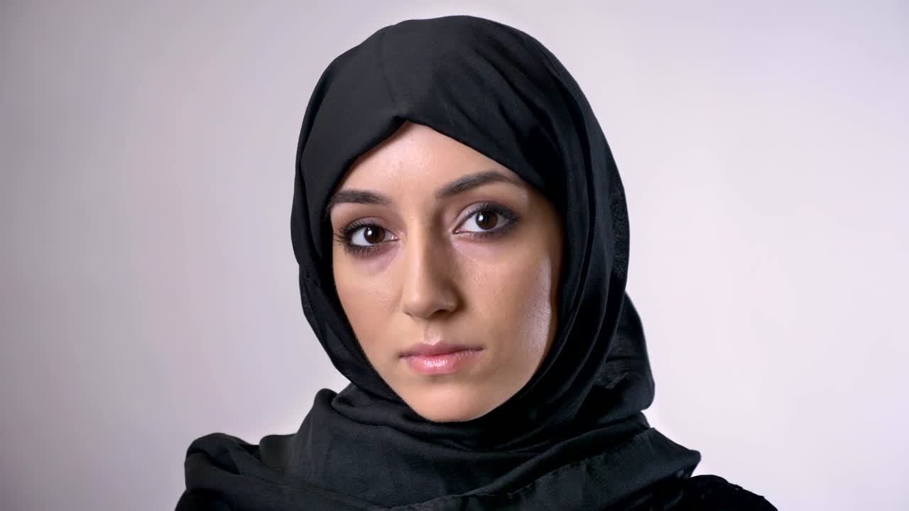 Young serious muslim girl in hijab is watching at camera, grey background