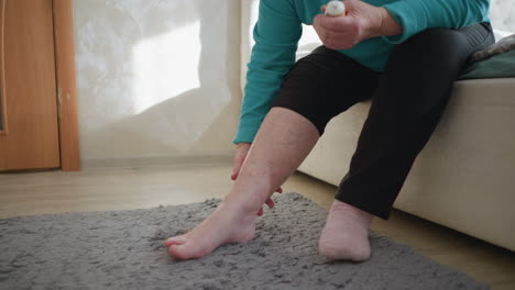 old woman in green applying ointment to her ankle while sitting on bed. one sock on, the other off, in a cozy bedroom setting. soft natural light fills the room, creating a calm, peaceful atmosphere