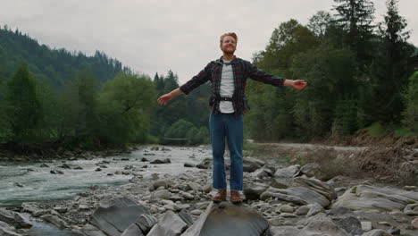 guy standing on rock at river shore. male tourist with backpack raising hands